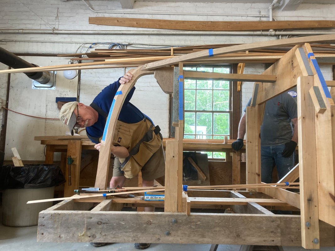 Bob Fuller leaning on the keel of a wooden boat frame to teach his students how to build a wooden boat in a shop and powered by south shore boatworks
