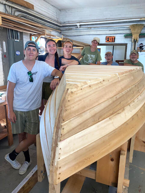 Students standing in a shop with the wooden boat they built at Bob Fullers class powered by South Shore Boatworks