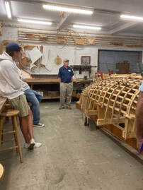 Bob Fuller and his students standing next to a framed wooden boat for their class on wooden boat building in a shop and presented by south shore boatworks