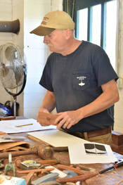 Bob Fuller of South Shore Boatworks standing over workbench with boat plans and books on it as he prepares to teach a wooden boat building class
