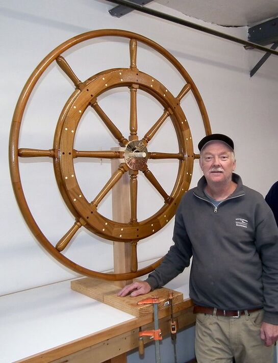 Bob Fuller standing next to very large wooden yacht steering wheel with bronze engraved hub and glossy varnished finish at the shop for south shore boatworks